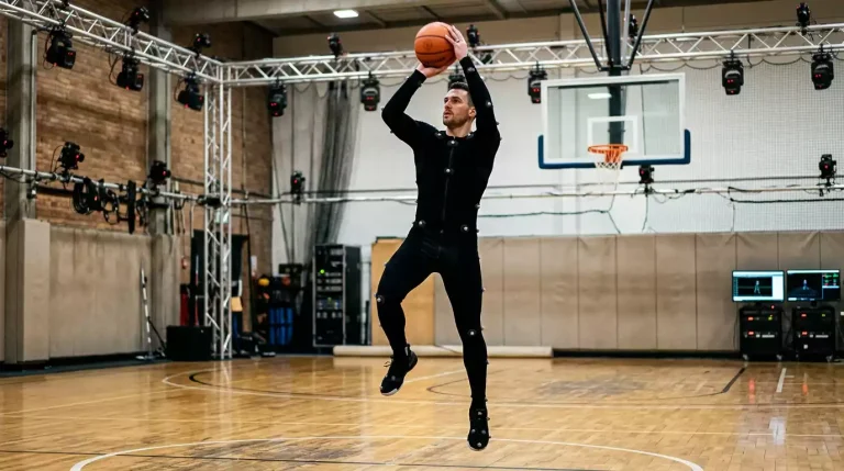 A motion-capture studio with a basketball player wearing a black motion-capture suit covered in reflective markers, performing a jump shot under arrays of tracking cameras mounted on the ceiling, on a small indoor practice court.