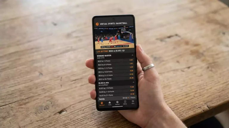 Smartphone displaying a virtual basketball winning margin market with a list of margin bands like '1-5', '6-10', '11-15' priced with decimal odds, held over a desk with the basketball preview visible.